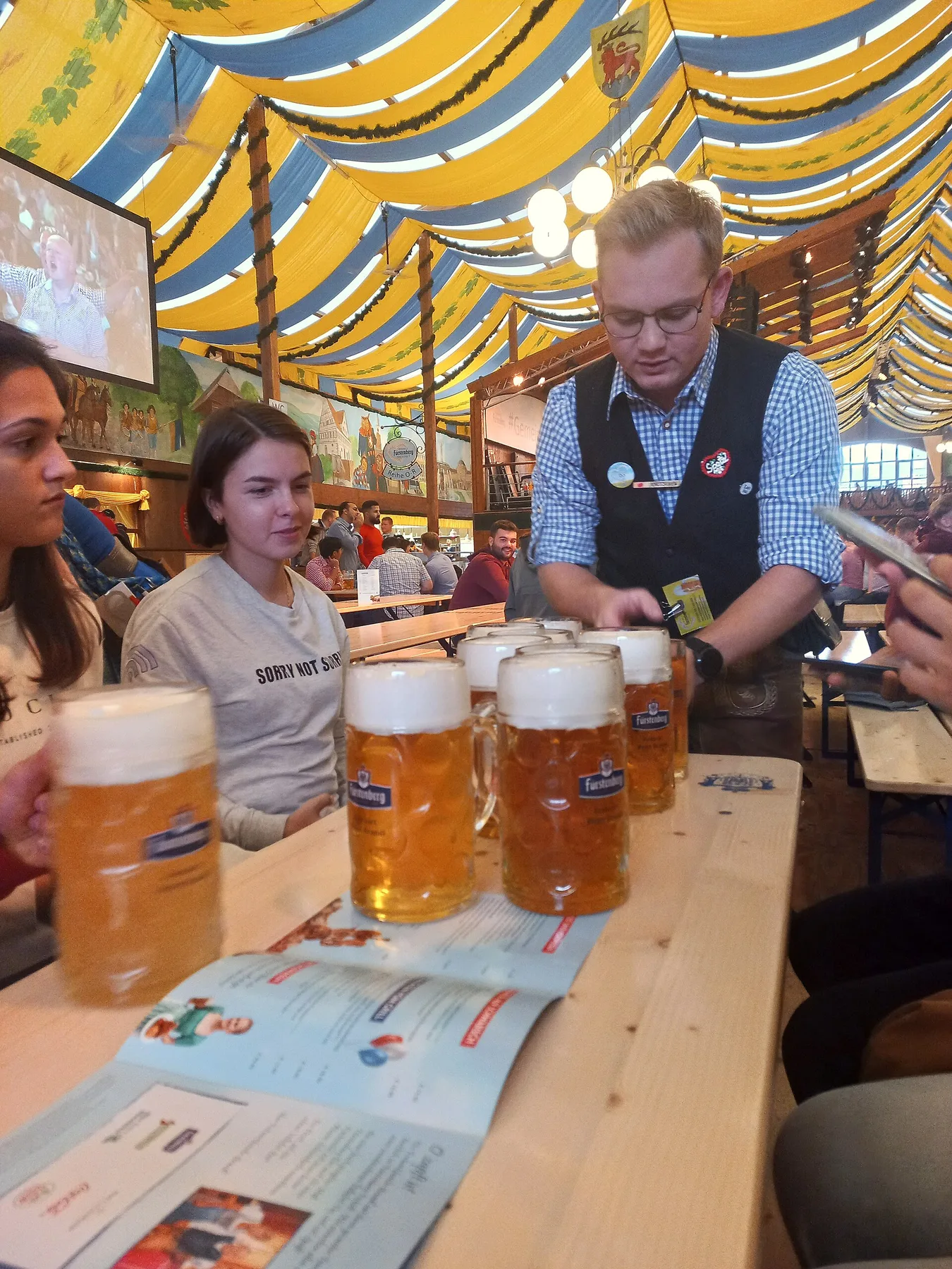 Server carrying several full glass beer mugs to a shared table inside a decorated festival tent